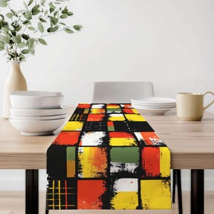 May include: A colorful table runner with an abstract design. The runner features a pattern of overlapping squares in red, yellow, green, and white against a black background. The design has a distressed, painted look, adding a modern touch to the table setting.