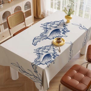 May include: A white tablecloth with a large blue seashell design. The tablecloth is on a wooden table with brown chairs. A small yellow plate with a dessert and a fork are on the table. A yellow vase with flowers is also on the table.