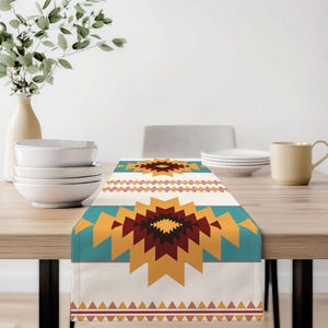May include: A rectangular table runner with a geometric pattern in shades of cream, teal, gold, and burgundy. The design features a central starburst motif and a border of triangles. The runner is placed on a wooden table, with tableware and a vase in the background.