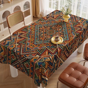 Op de afbeelding: Een rechthoekig tafelkleed met een levendig geometrisch patroon in tinten oranje, blauw, groen en bruin. Het ontwerp bevat driehoeken en vierkanten, waardoor een gedurfde, kleurrijke uitstraling ontstaat voor een eettafel. Een gele bord met een kleine cake staat op de tafel.
