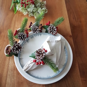 May include: A festive table setting featuring white plates with a blue rim, a folded linen napkin, and decorative pine cone napkin rings. The rings are adorned with faux evergreen sprigs, red berries, and pine cones, creating a holiday theme.