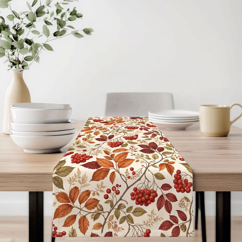 May include: A rectangular table runner featuring a repeating botanical pattern of autumn-colored leaves in shades of orange, brown, and red, interspersed with clusters of small red berries on a cream-colored background, laid flat on a wooden table.