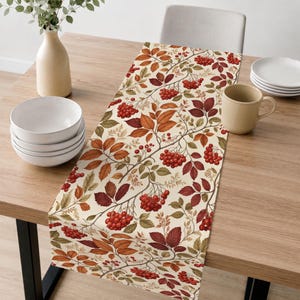 May include: A rectangular table runner featuring a cream-colored background with a repeating pattern of autumn-toned leaves in shades of orange, brown, and green, interspersed with clusters of small, vibrant red berries across the fabric surface.