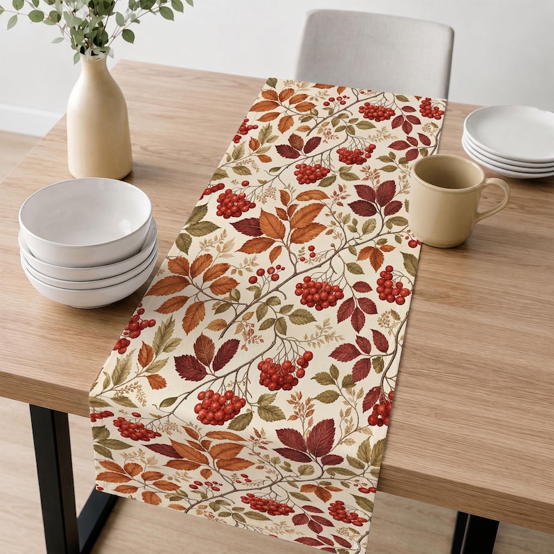 May include: A rectangular table runner featuring a cream-colored background with a repeating pattern of autumn-toned leaves in shades of orange, brown, and green, interspersed with clusters of small, vibrant red berries across the fabric surface.