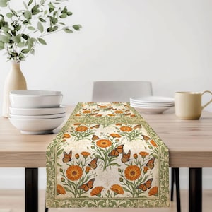 May include: A rectangular table runner featuring a repeating pattern of orange marigold flowers and monarch butterflies on a cream background, framed by a decorative sage green border with intricate scrollwork and floral motifs.