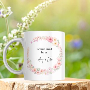 May include: White ceramic mug with a floral wreath design in pink and cream colors. The mug has the text "Always loved by us" and the names "Amy & Luke". The mug is sitting on a wooden surface.