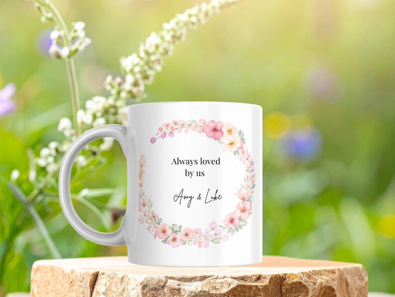 May include: White ceramic mug with a floral wreath design in pink and cream colors. The mug has the text "Always loved by us" and the names "Amy & Luke". The mug is sitting on a wooden surface.