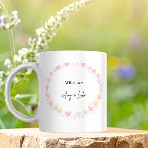 May include: White ceramic mug with a handle, featuring a heart-shaped wreath in pastel colors. The text "With Love" and names "Amy & Luke" are centered within the wreath. The mug is set against a blurred green and yellow background.