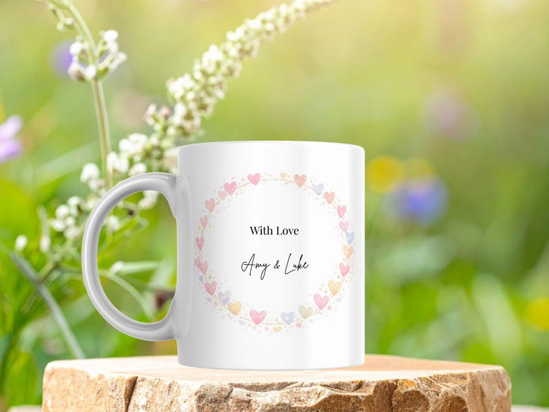 May include: White ceramic mug with a handle, featuring a heart-shaped wreath in pastel colors. The text "With Love" and names "Amy & Luke" are centered within the wreath. The mug is set against a blurred green and yellow background.