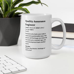 Peut inclure: Une tasse à café en céramique blanche avec une définition en noir et blanc d'un "Quality Assurance Engineer" imprimée sur le devant. La définition comprend la prononciation phonétique du mot "engineer" et une description humoristique du travail.