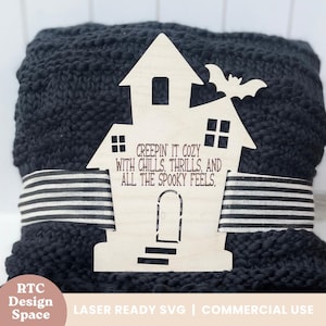 Pode incluir: Decoração de casa assombrada em madeira com um recorte de morcego, texto "CREEPIN IT COZY WITH CHILLS, THRILLS, AND ALL THE SPOOKY FEELS". A decoração é colocada sobre um cobertor cinza escuro com uma fita listrada em preto e branco.