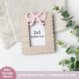 May include: A wooden photo frame with a scalloped edge and a pink bow at the top. The frame is designed for a 2x3 inch wallet-sized photo. The text "2x3 wallet size" is visible within the frame.