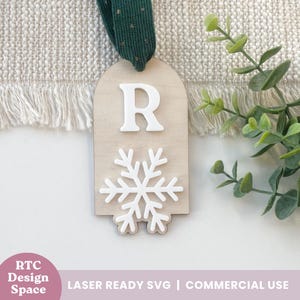 Puede incluir: Adorno navideño de madera con un copo de nieve blanco y la letra "R". El adorno tiene una cinta verde y el texto "RTC Design Space" y "LASER READY SVG | COMMERCIAL USE".