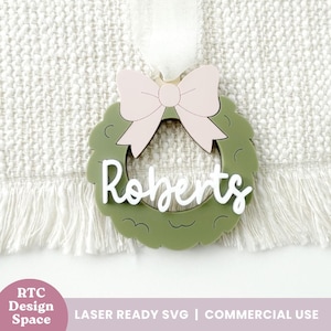 Peut inclure: Ornement de Noël personnalisé en forme de couronne verte avec un nœud rose et le nom "Roberts" en écriture blanche. L'ornement est suspendu à un ruban blanc. Le design est découpé au laser.
