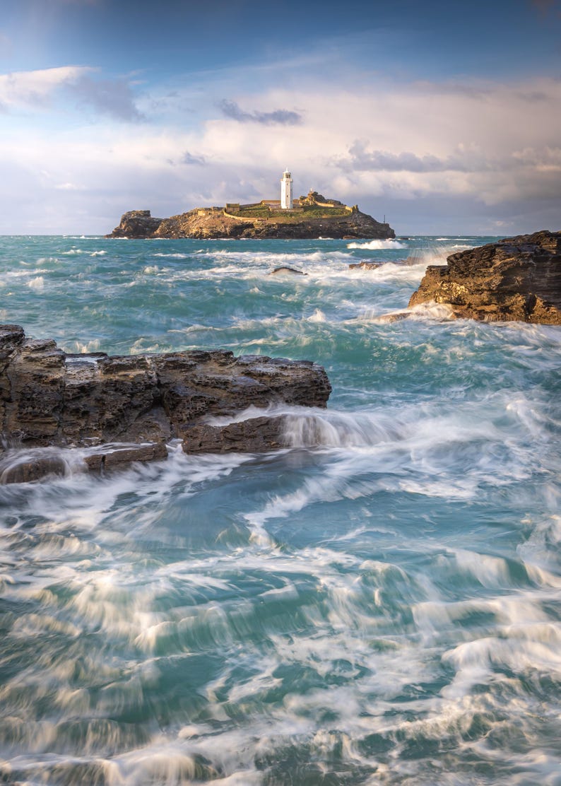 K&ouml;nnte beinhalten: Ein wei&szlig;er Leuchtturm steht auf einer felsigen Insel, umgeben von turbulenten t&uuml;rkisfarbenen Meereswellen, die gegen dunkle, zerkl&uuml;ftete K&uuml;stenfelsen prallen, unter einem wolkenverhangenen Himmel im Licht des sp&auml;ten Nachmittags.