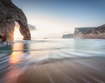 Könnte beinhalten: Landschaftsfotografie einer Küste bei Sonnenuntergang. Ein natürlicher Felsbogen steht im Meer, mit der Sonne, die durch ihn scheint. Das Wasser ist eine Mischung aus Blau und Weiß, und der Strand ist hellbraun. Der Himmel ist eine Mischung aus Blau und Orange.