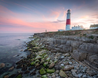 Könnte beinhalten: Ein weißer und roter Leuchtturm steht auf einer felsigen Klippe mit Blick auf ein ruhiges Meer. Der Himmel ist in sanftem Rosa und Lila gefärbt und erzeugt einen wunderschönen Sonnenuntergang.
