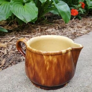 May include: A brown and yellow glazed ceramic pitcher with a handle. The pitcher has a rustic, vintage look.
