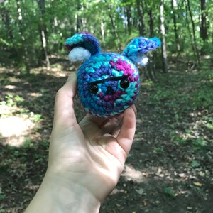 May include: A crocheted stuffed animal with a blue, purple, and pink color scheme. The animal has a round body with two floppy ears and black eyes.