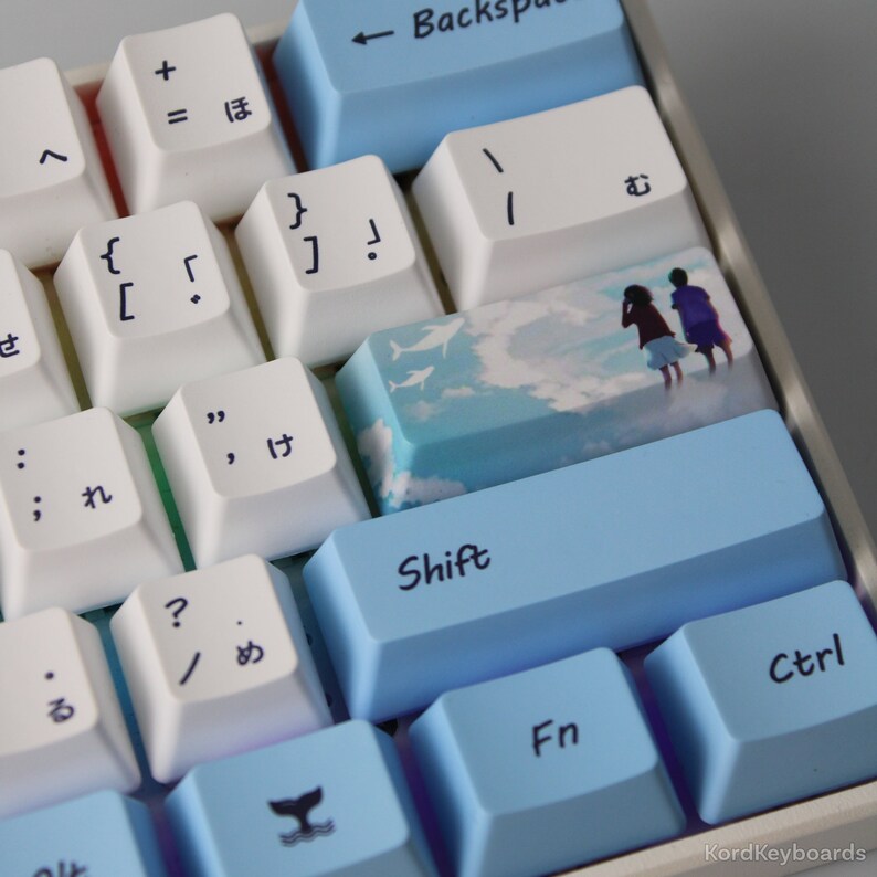 May include: A close-up of a mechanical keyboard with custom keycaps. The keyboard has white and light blue keycaps with Japanese characters and symbols. One keycap features an image of two people on a beach. The "Shift" key is light blue.