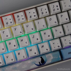 May include: A white mechanical keyboard with Japanese characters and English letters, with a rainbow backlight. The keycaps are white. One keycap has a silhouette of a person and a cat on a crescent moon. The keyboard is a modern design.