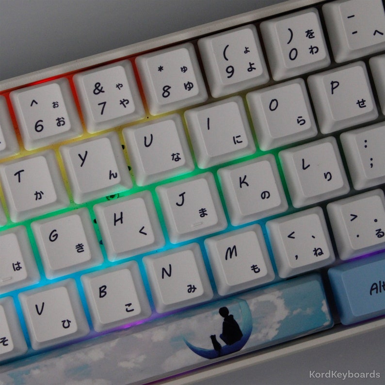 May include: A white mechanical keyboard with Japanese characters and English letters, with a rainbow backlight. The keycaps are white. One keycap has a silhouette of a person and a cat on a crescent moon. The keyboard is a modern design.