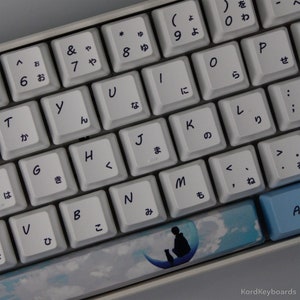 May include: A white mechanical keyboard with Japanese hiragana characters and English letters. The space bar has a blue and white design with a silhouette of a person and a cat on a crescent moon.