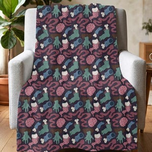 May include: A dark purple throw blanket featuring a repeating pattern of severed blue and green hands, pink brains, white eyeballs, and white bones. The graphic print is distributed evenly across the fabric surface in a consistent, repeating design.