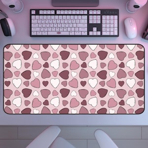May include: A rectangular desk mat featuring a repeating pattern of hearts in various shades of pink, mauve, and white on a light pink background. The mat has a black border and is positioned below a computer keyboard, a white mouse, and headphones.