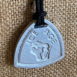 May include: A silver-colored pendant with the words "ICE AGE" and a mammoth graphic. The pendant is triangular and hangs from a black cord. The background is a textured, tan fabric.