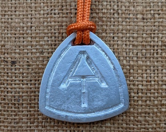 May include: A silver-colored pendant with the Appalachian Trail logo, a white triangle with a vertical line through it. The pendant is attached to an orange cord. The background is a textured, brown surface.