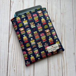 May include: A navy blue fabric sleeve for an e-reader featuring a repeating pattern of colorful stacked books, small mugs, and green grapes. A white rectangular label with the text Darula Designs Handmade is stitched onto the lower right corner.