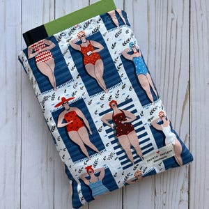 May include: A blue book cover with a white background and a pattern of women in swimsuits. The women are all wearing red, blue, or white swimsuits and have red lips and sunglasses. The text "Darula Designs Handmade" is on the bottom right corner of the cover.