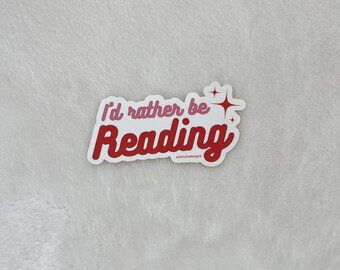 May include: A white sticker with red and pink text that reads "I'd rather be Reading" with a sparkle design. The sticker has the text "adamjade.designs" in small letters at the bottom.
