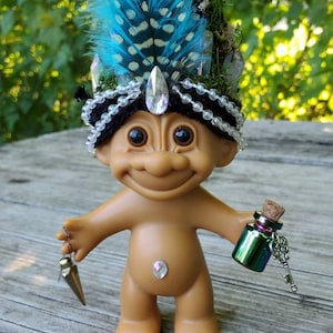May include: A small, brown troll doll with black hair wearing a crown made of crystals, feathers, and beads. The troll is holding a small, green glass bottle with a key attached.
