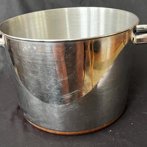 May include: A stainless steel stockpot with a polished finish and two side handles. The exterior walls are reflective, showing light reflections, and the base features a distinct copper-coloured band around the bottom edge of the cooking vessel.