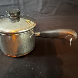 May include: A small, stainless steel saucepan with a lid and a dark wooden handle. The handle is attached to the side of the pot with a metal bracket. The lid has a black knob. The pot is sitting on a dark surface.
