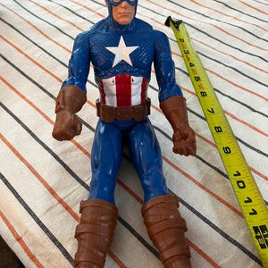 May include: A blue and red plastic action figure of Captain America wearing a blue and white costume with a red, white, and blue star on the chest. The figure is wearing brown boots and has a red, white, and blue belt.