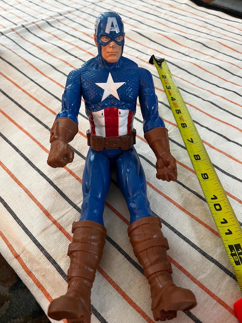 May include: A blue and red plastic action figure of Captain America wearing a blue and white costume with a red, white, and blue star on the chest. The figure is wearing brown boots and has a red, white, and blue belt.