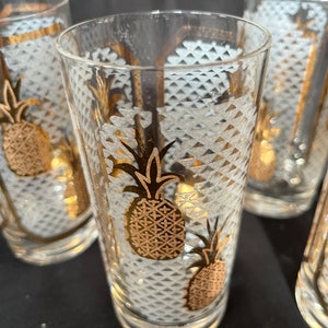 Może przedstawiać: Cztery przezroczyste szklanki z wzorem białego i złotego ananasa. Szklanki mają fakturowaną powierzchnię i idealnie nadają się do serwowania koktajli lub soku.