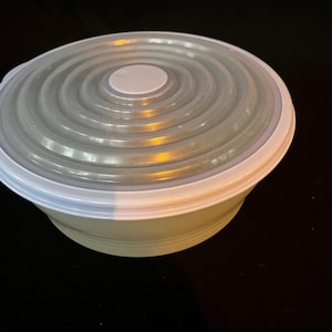 May include: A round, light green bowl with a white rim and a gray, ridged lid. The lid has a small, white circular handle in the center. The bowl is likely made of plastic and is designed for food storage.