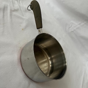May include: A stainless steel measuring cup with a dark brown handle and a small metal ring. The cup has a rounded shape and a red rim. The handle is curved and attached to the cup with a small metal piece.