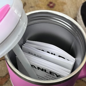 May include: A pink and silver insulated travel mug with a white lid and straw. Inside the mug are several white instruction manuals. The word "STANLEY" is printed on the side of the mug.