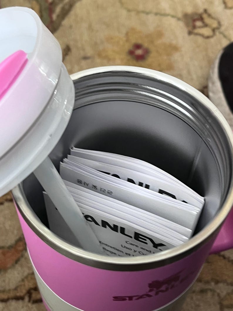 May include: A pink and silver insulated travel mug with a white lid and straw. Inside the mug are several white instruction manuals. The word "STANLEY" is printed on the side of the mug.