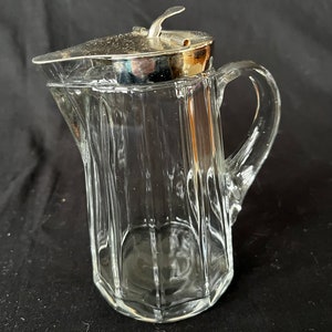 May include: A clear glass pitcher with a silver lid and a handle. The pitcher has a faceted design.
