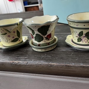 Może przedstawiać: Trzy małe ceramiczne doniczki z zielonymi i żółtymi wzorami kwiatowymi. Doniczki mają pasujące podstawki.