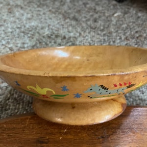 May include: A light brown wooden bowl with a pedestal base. The bowl is decorated with painted floral and animal designs in blue, yellow, and red. The bowl is sitting on a wooden surface.