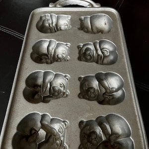 Może przedstawiać: Antyczna forma do pieczenia z żeliwa z ośmioma foremkami w kształcie niedźwiedzia. Forma jest srebrna i ma uchwyt.