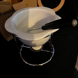 Puede incluir: Olla de fondue de cerámica blanca con un soporte cromado y un tenedor de fondue negro. La olla está sobre una superficie negra.