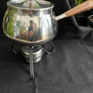 Puede incluir: Una olla de fondue de plata con un mango y una tapa de madera, sentada sobre un soporte de metal negro.